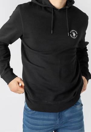 Mężczyzna dopasowujący czarną bluzę z kapturem U.S. Polo Assn. z logo na piersi, noszący niebieskie dżinsy, przycięte przy szyi i biodrach na białym tle.