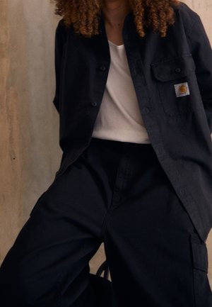 Personne aux cheveux bouclés portant une chemise noire à boutons avec le logo Carhartt sur un t-shirt blanc et un pantalon cargo noir, posant devant un mur beige.