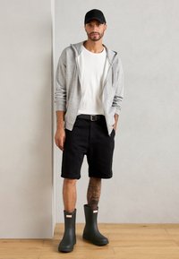 Sweat à zip gris, t-shirt blanc, shorts en denim noirs et bottes en caoutchouc vertes avec des accents rouges. Sol en bois et fond neutre.