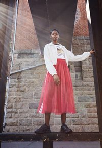 Modell iklädd en vit cropped bomberjacka över en grafisk t-shirt, med en genomskinlig röd midi kjol och svarta sneakers. Nätstrumpbyxor synliga.