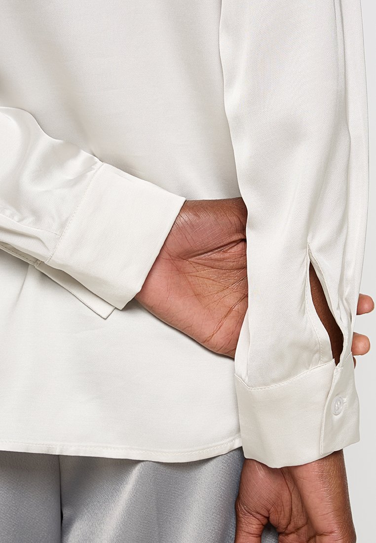 Langärmliges beiges Hemd mit glatter Satin-Oberfläche, ausgestattet mit einem geknöpften Bündchen und einem Schlitzdetail am Ärmelabschluss.