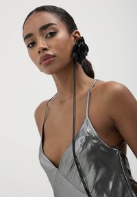 Metallisch silbernes Kleid mit tiefem Ausschnitt und dünnen Trägern. Schwarzes Stoffblumenaccessoire im Haar. Glatte Textur insgesamt.