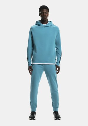 Man standing wearing light blue hoodie and matching joggers with black sneakers, facing forward on white background.
