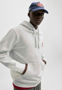 Vaaleanharmaa huppari etutaskulla, nyörisäädettävällä hupulla ja pyöreällä kirjotulla logolla. Yhdistetty punaiseen paitaan alla ja harmaisiin housuihin.