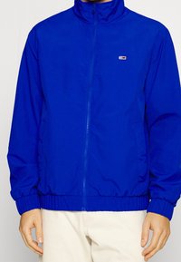 Blue zip-up jacket with a high collar, elastic cuffs, and hem. Features a small logo on the left chest. Made from a smooth, lightweight material.