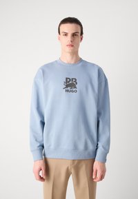 Sudadera verde claro de cuello redondo con puños y dobladillo acanalados, que presenta una impresión gráfica negra de un toro y el texto "Hugo" en la parte delantera.