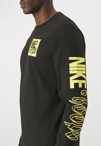 Maglietta nera a maniche lunghe in cotone con un logo grafico giallo brillante sul davanti e sulla manica, caratterizzata da un design a collo rotondo standard.