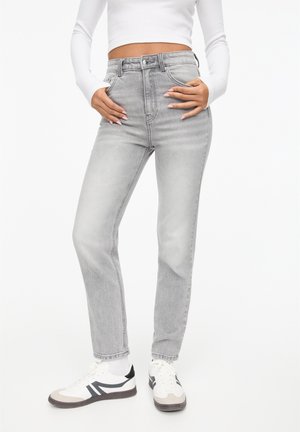 Person wearing high-waist faded gray jeans, white sneakers, and a white long-sleeve cropped top, standing with hands near pockets.