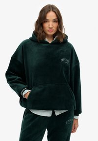 Haut en velours vert avec une capuche spacieuse, une poche frontale et un logo brodé "SUPERDRY". Comprend des poignets côtelés et une coupe décontractée.