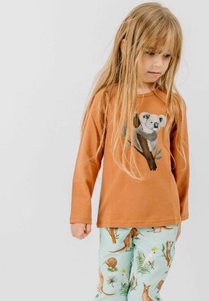 Young girl with long blonde hair wearing a brown top with a koala print and light blue pants featuring various animals and plants.