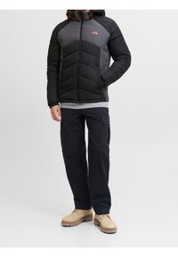 Mężczyzna ubrany w czarno-szarą pikowaną kurtkę, czarne spodnie typu cargo i beżowe buty, stojący na białym tle.