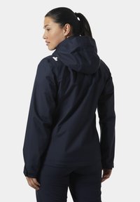 Marineblauwe waterdichte jas met capuchon, voorzien van een strak ontwerp, verstelbare manchetten en een reflecterend logo op de schouder. Soepele textuur.