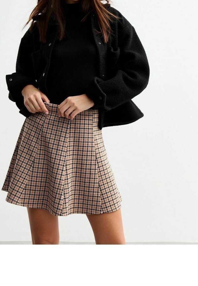 Femme portant une veste noire et un haut noir avec une jupe plissée à carreaux beige et noir, ajustant la taille avec les deux mains.