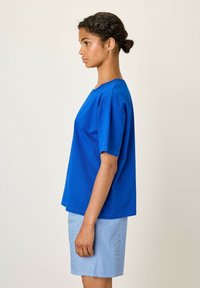 Mėlyna medvilninė marškinėliai su trumpomis rankovėmis ir laisvu kirpimu, derinami su mėlynomis dryžuotomis šortais. Audinys atrodo lygus ir lengvas.