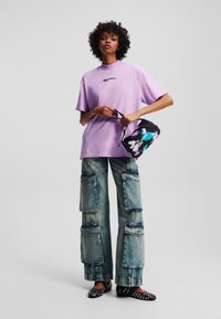 Lilla oversized t-skjorte med logo, vide cargo-bukser med flere lommer, svarte flate sko med nagler, modell som står mot en grå bakgrunn.