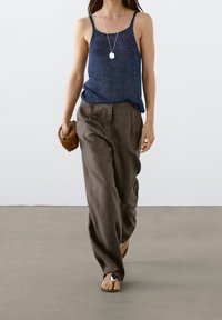 Marineblauwe gebreide tanktop met dunne bandjes, gecombineerd met losse bruine broeken. Sandalen met een ronde accent. Model houdt een geweven bruine clutch vast.