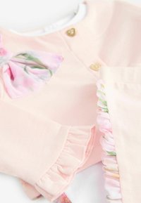 Helles, pinkes Langarmshirt mit floralem Schleifenakzent, goldenen Herzknöpfen und Rüschenärmeln. Baumwollmaterial mit einer glatten Textur.