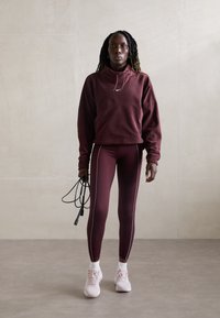 Bordeauxfarbener Kapuzenpullover und Leggings mit weißen Seitenstreifen, kombiniert mit weißen Knöchelsocken und hellrosafarbenen Sportschuhen; hält ein Springseil.