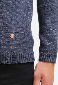 Mörkblå stickad tröja med ribbade muddar och nederkant, med en liten logotypplatta i brunt läder på framsidan.