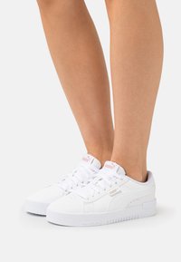 Weiße Sneaker mit einem glatten Lederobermaterial, runder Zehenpartie, flacher Sohle und auffälligen Schnürsenkeln. Enthält ein dezentes Logo und strukturierte Akzente.