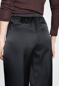 Pantaloni in satin nero con una texture liscia e vita elastica, dotati di due tasche posteriori. Il tessuto riflette la luce in modo delicato.
