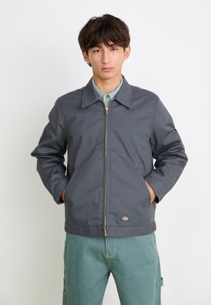Veste grise avec fermeture éclair, grand col, poches avant et patch logo ; portée par-dessus une tenue verte avec une chemise rayée.