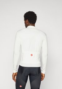 Mann in weißem langärmligen Fahrradtrikot und schwarzen gepolsterten Fahrradhosen, der mit dem Rücken zu einer schlichten Hintergrund steht.