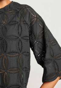 Blouse en tissu noir avec une grande broderie à œillets circulaires en motif floral, dotée de manches larges arrivant au coude et de bords festonnés.
