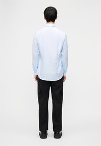 Άνδρας που στέκεται με την πλάτη γυρισμένη, φορούντας ανοιχτό γαλάζιο πουκάμισο με μακριά μανίκια, μαύρο παντελόνι cargo και μαύρα παπούτσια, μπροστά σε απλό γκρι φόντο.