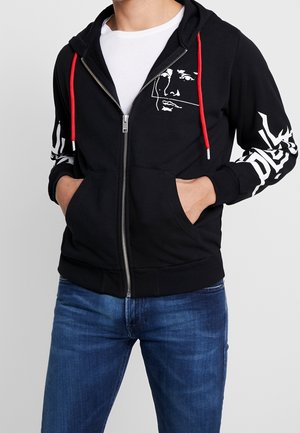 Homme portant un sweat à capuche noir zippé avec des cordons rouges, un visage graphique blanc sur la poitrine et des lettres blanches sur la manche, porté avec un jean bleu.