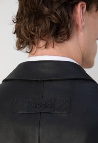 Czarna skórzana kurtka z wyciętym kołnierzem i markową metką z napisem "HUGO" na plecach. Gładka tekstura z minimalnymi detalami szycia.
