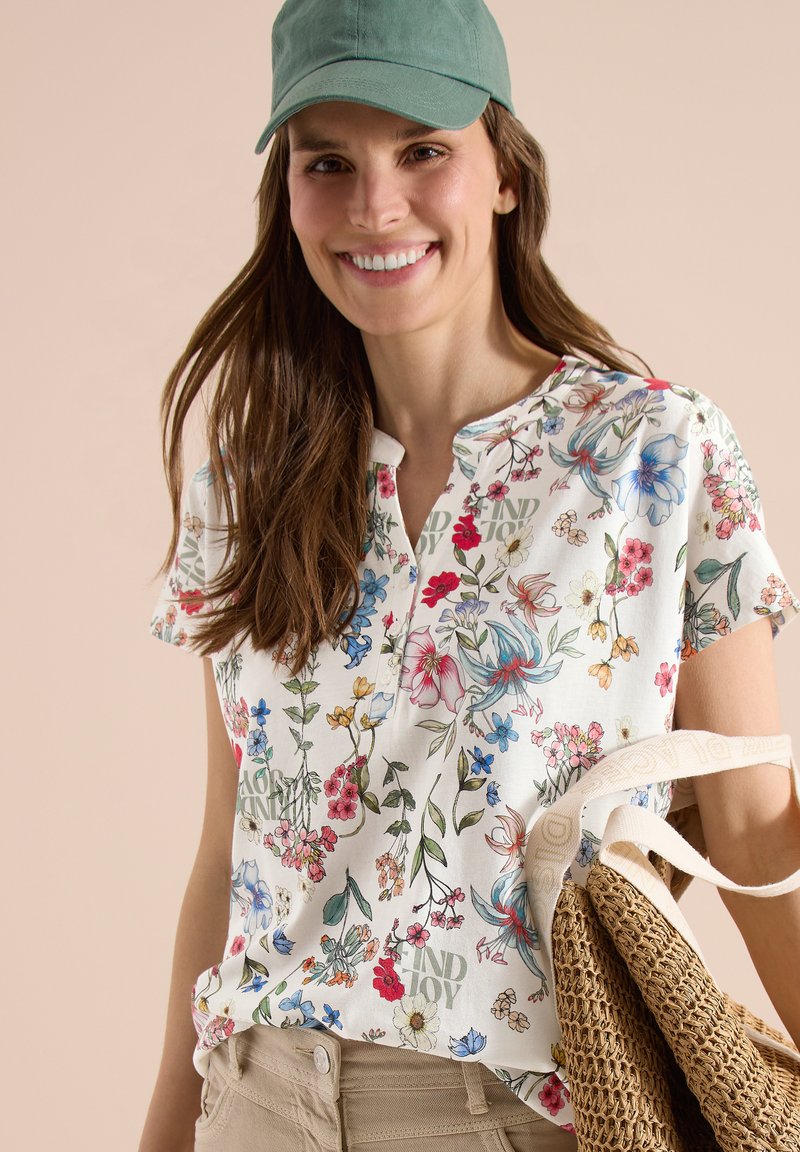 Smilende kvinde iført grøn kasket, blomstret kortærmet skjorte, beige bukser, der holder vævet taske med cremefarvet rem mod beige baggrund.
