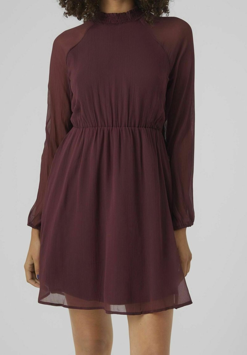 Femme portant une robe bordeaux à manches longues, col montant, longueur au genou, taille cintrée et tissu transparent aux manches et à la jupe.