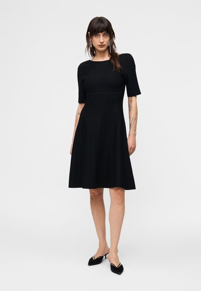 Γυναίκα που φοράει μαύρο φόρεμα μέχρι το γόνατο με μανίκια μέχρι τον αγκώνα, στέκεται όρθια με μαύρα μυτερά παπούτσια slingback μπροστά σε απλό φόντο.