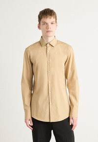 Cămașă lungă, de culoare maro deschis, cu guler ascuțit și nasturi la front. Materialul pare neted, cu un ușor luciu. Asortată cu pantaloni negri.