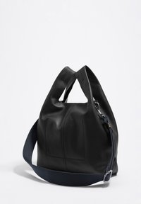 Črna usnjena torba z mehkimi, teksturiranimi površinami, okroglim oblikovanjem in mornarsko modrim platnenim naramnicam. Ima zadrgo in vidne šive.