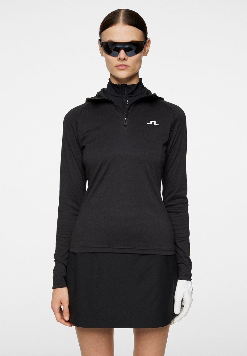 Svart långärmad zip-up topp med hög hals, med en vit logotyp på bröstet. Bärs med en svart kjol och vit handske.