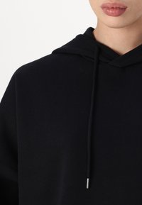 Μαύρο φούτερ με κουκούλα και υφή υφάσματος. Διαθέτει τσέπη τύπου καγκουρό, κορδόνι με μεταλλικές άκρες και σχέδιο άνετης εφαρμογής.