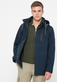 Νεαρός άνδρας φορώντας ναυτικό μπλέ φούτερ με κουκούλα πάνω από πράσινο πουλόβερ με μισό φερμουάρ και μπεζ παντελόνι cargo, στέκεται με το ένα χέρι στην τσέπη μπροστά από λευκό φόντο.