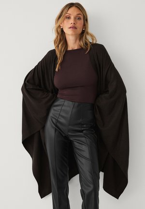 Bruine kimono-stijl wikkel gemaakt van zachte stof, met brede mouwen en een open voorkant, gecombineerd met hoge zwarte leren broeken.