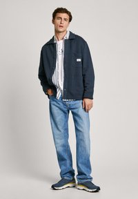 Marininspirerad knapplångjacka med två frontfickor, lager på en vit randig skjorta, tillsammans med ljusblå jeans och mörka träningsskor.