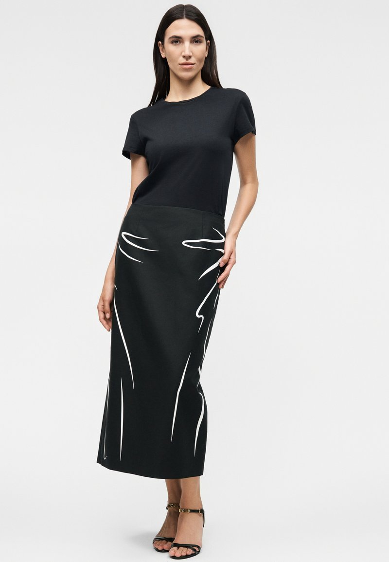 Woman wearing a black short-sleeve top paired with a black midi skirt featuring white abstract line designs, and black open-toe heels.