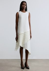 Rochie albă fără mâneci, cu tiv asimetric, prevăzută cu franjuri texturate în partea de jos. Material neted, cu decolteu drept și design minimalist.