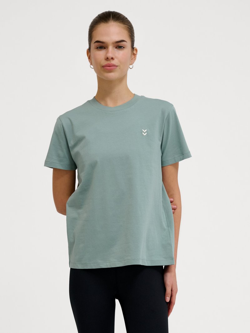 Junge Frau mit heller Haut und zurückgebundenen Haaren, die ein hellgrünes kurzärmeliges T-Shirt und schwarze Hose vor weißem Hintergrund trägt.
