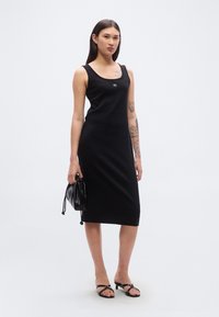 Μαύρο αμάνικο φόρεμα από απαλό ύφασμα, με εφαρμοστή σιλουέτα και διακριτική λεπτομέρεια λογότυπου. Φορεμένο με μαύρα σανδάλια και μικρή τσάντα.
