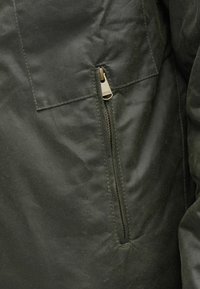 Jachetă verde închis cu o buzunar cu fermoar vizibil și un capăt de tab bronzat. Țesătura are o textură netedă, mată, cu un ușor luciu.