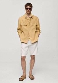 Světle žlutá košile na knoflíky s dvěma náprsními kapsami, v kombinaci s bílými šortkami a hnědými sandály, nošena s tmavými slunečními brýlemi.