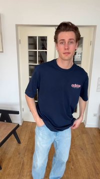 Νέος άνδρας που φοράει μπλε σκούρο Adidas Team t-shirt και ανοιχτό μπλε τζιν, στέκεται σε ένα δωμάτιο με ξύλινο πάτωμα και πόρτες με τζάμι.