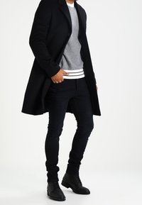 Czarny długi płaszcz na szarym dzianinowym swetrze z białymi paskami na dole, w połączeniu z ciemnymi dopasowanymi jeansami i czarnymi skórzanymi butami.