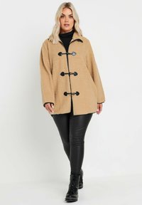 Beige faux shearling takki rento leikkauksella, mustat solkipainikkeet, pyöreä kaulus ja pitkät hihat. Vaatetus mustien keinonahkahousujen päällä.